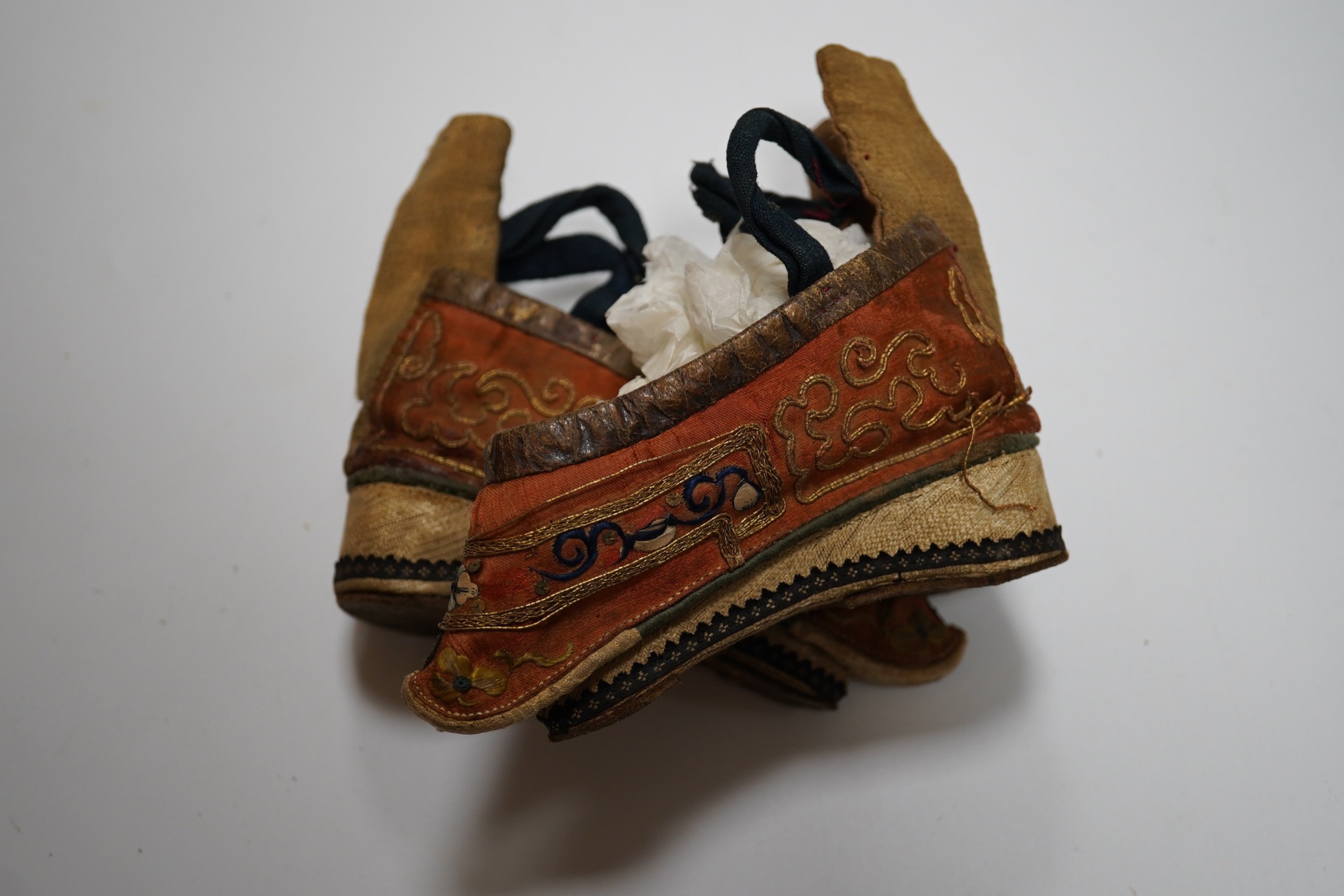 A pair of Chinese silk and gold thread embroidered ladies shoes, with leather binding around the top and gold brocade platform heels with fine blue braiding, 12cm long. Condition - good for age and use